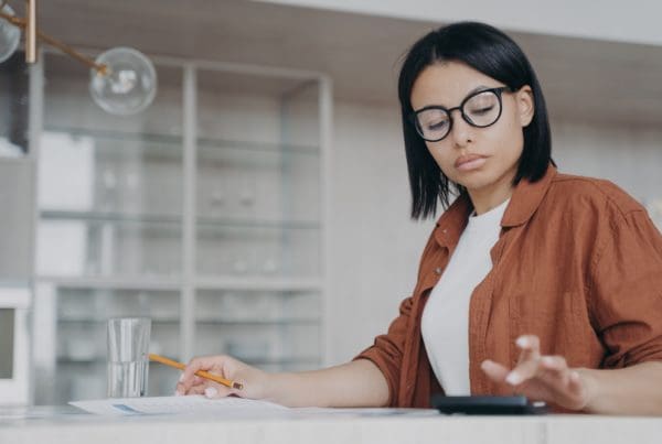 Focused female in glasses counts expenses on calculator, managing project budget, sitting at desk